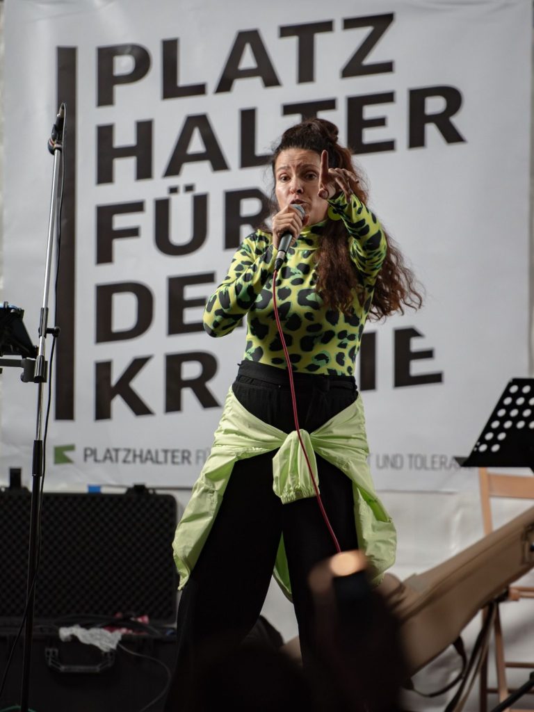 Sängerin RiA REiSER mit Mikrofon auf der Bühne vor Banner ‚PLATZ HALTER FÜR DEMOKRATIE‘.