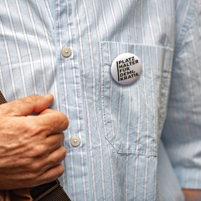 Nahaufnahme eines Hemds mit einer Brusttasche, an der ein runder Button mit der Aufschrift 'PLATZ HALTER FÜR DEMOKRATIE' befestigt ist